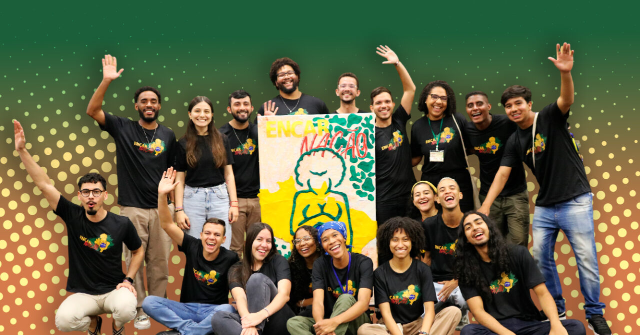 Grupo de jovens reunido e sorridente, vestindo camisetas pretas do evento Encarnação. Ao centro, seguram um painel colorido com ilustração e a palavra “Encarnação”. O fundo apresenta tons de verde e dourado com elementos gráficos pontilhados.