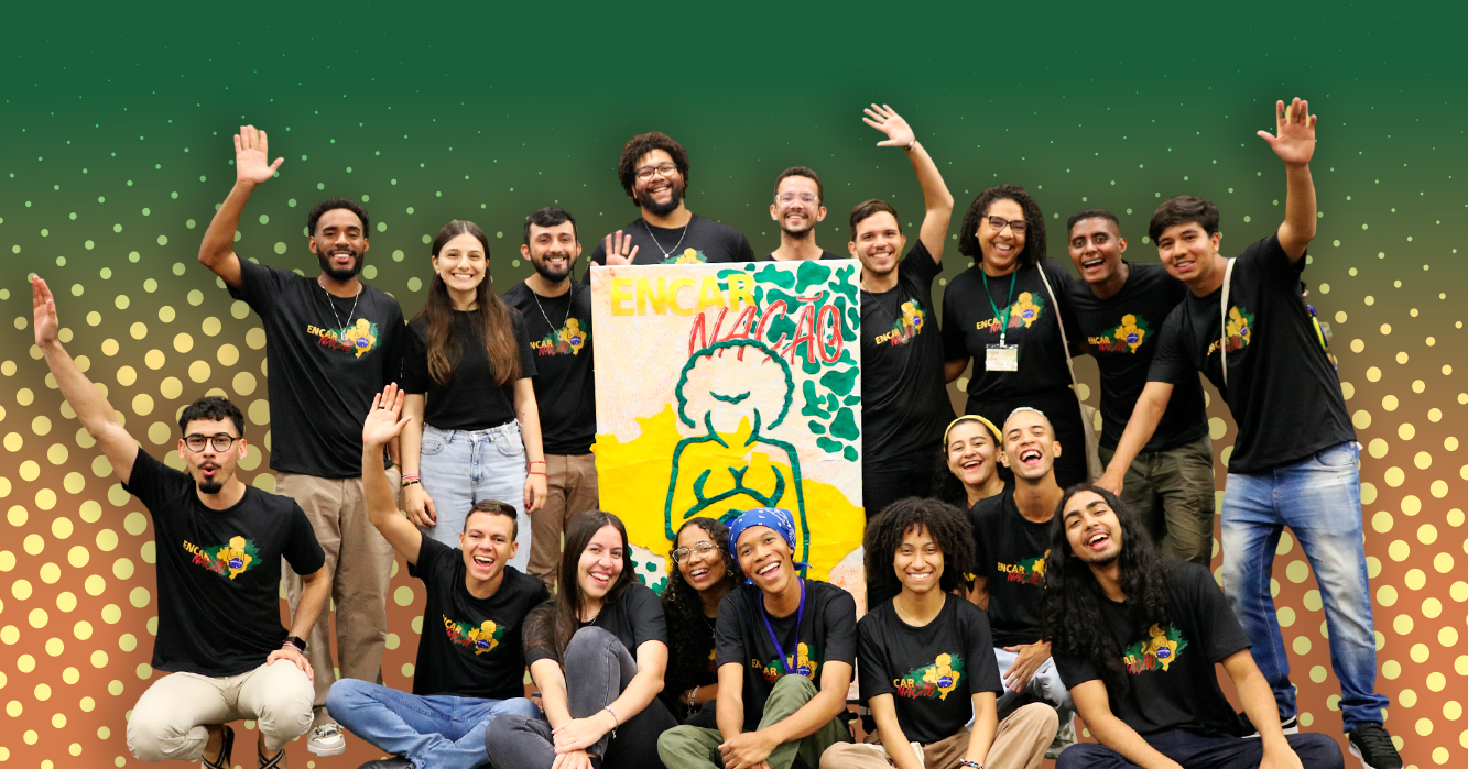 Grupo de jovens reunido e sorridente, vestindo camisetas pretas do evento Encarnação. Ao centro, seguram um painel colorido com ilustração e a palavra “Encarnação”. O fundo apresenta tons de verde e dourado com elementos gráficos pontilhados.