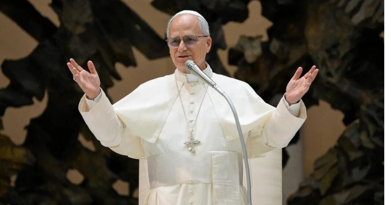 Papa Leão XIV, vestido de branco, fala ao microfone com os braços abertos durante uma audiência no Vaticano.