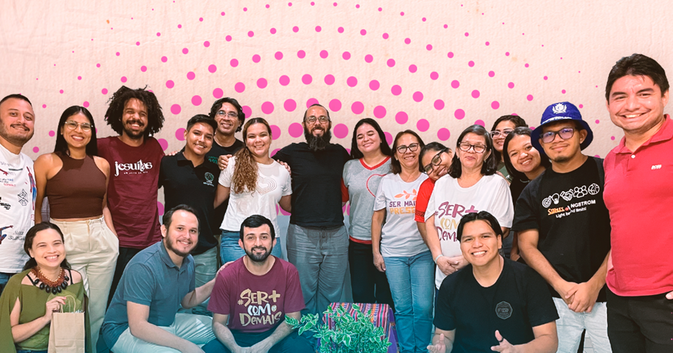 Grupo de jovens e adultos reunidos para uma foto durante atividade formativa, em ambiente interno, com painel rosa ao fundo.