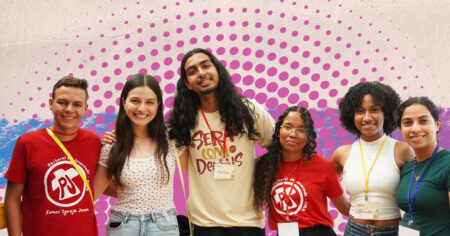 Grupo de seis jovens reunidos e sorrindo diante de um fundo gráfico em tons de rosa, azul e roxo. Alguns usam camisetas da Pastoral da Juventude e crachás de identificação.
