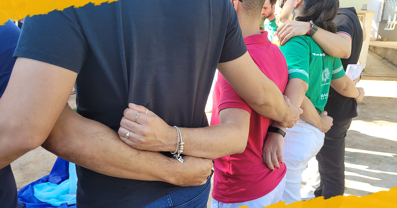 Jovens participam de uma dinâmica em grupo, formando uma corrente com os braços entrelaçados. A cena transmite união, acolhimento, convivência e espírito de comunidade. Ao fundo, uma participante veste camiseta verde do MAGIS Brasil.