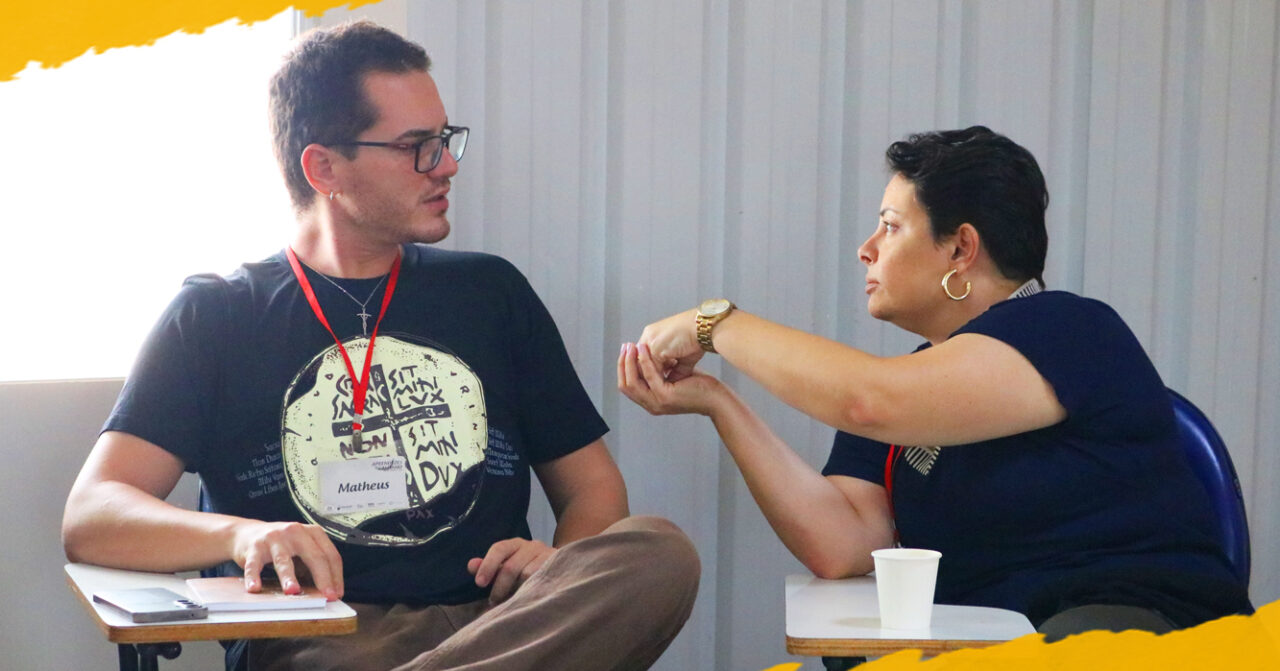 Homem e mulher conversam sentados frente a frente durante um encontro. Ele usa camiseta preta, óculos e crachá; ela veste blusa azul-escura e gesticula com as mãos enquanto fala.