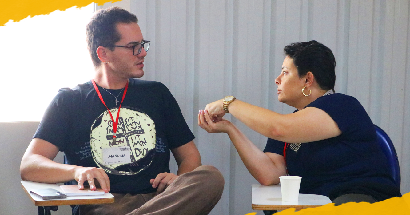 Homem e mulher conversam sentados frente a frente durante um encontro. Ele usa camiseta preta, óculos e crachá; ela veste blusa azul-escura e gesticula com as mãos enquanto fala.