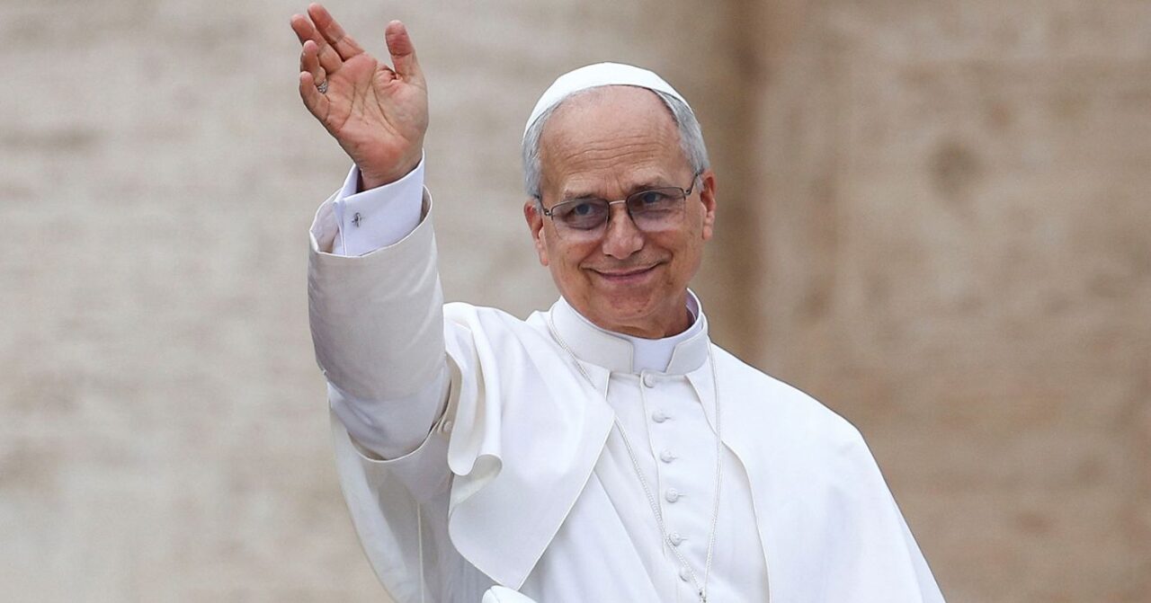 Papa Leão XIV, vestido de branco, sorri e acena com a mão direita durante aparição pública.