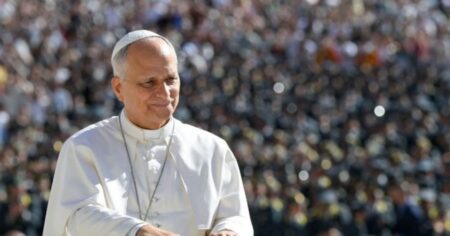 Papa Leão XIV, vestido de branco, caminha sorrindo diante de uma grande multidão reunida na Praça de São Pedro, no Vaticano.