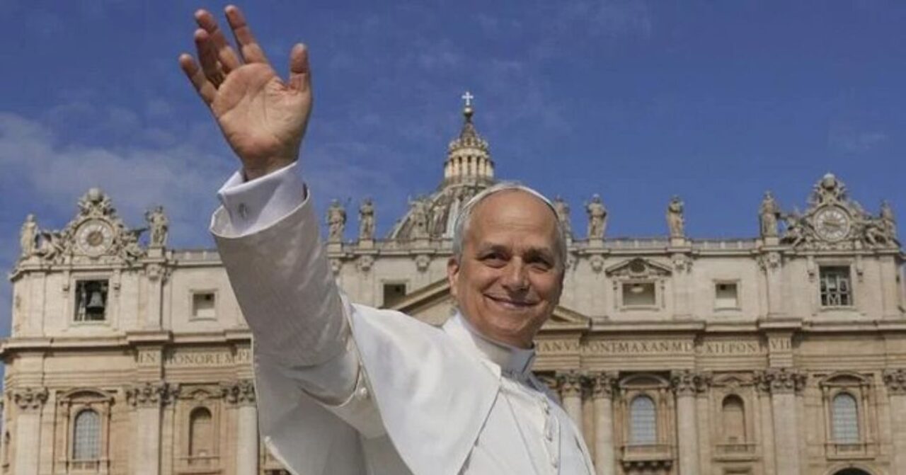 Papa Leão XIV acena sorrindo diante da Basílica de São Pedro, no Vaticano, sob céu azul.