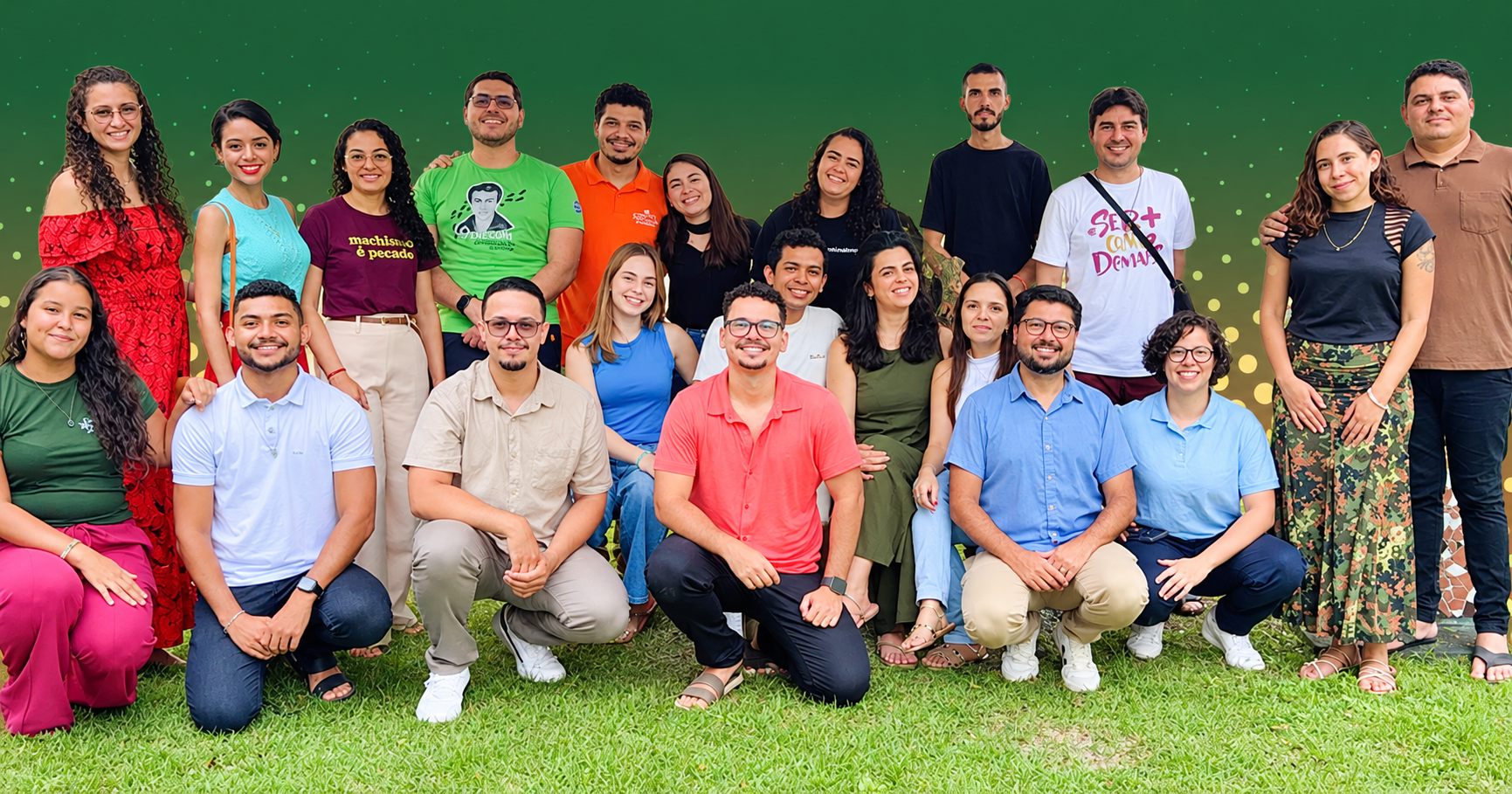 Grupo de jovens e adultos posa para foto em duas fileiras, alguns em pé e outros agachados, sobre um gramado e diante de um fundo verde.