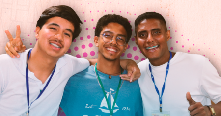 Três jovens sorridentes e abraçados demonstram amizade e companheirismo, diante de um fundo claro com detalhes em rosa.