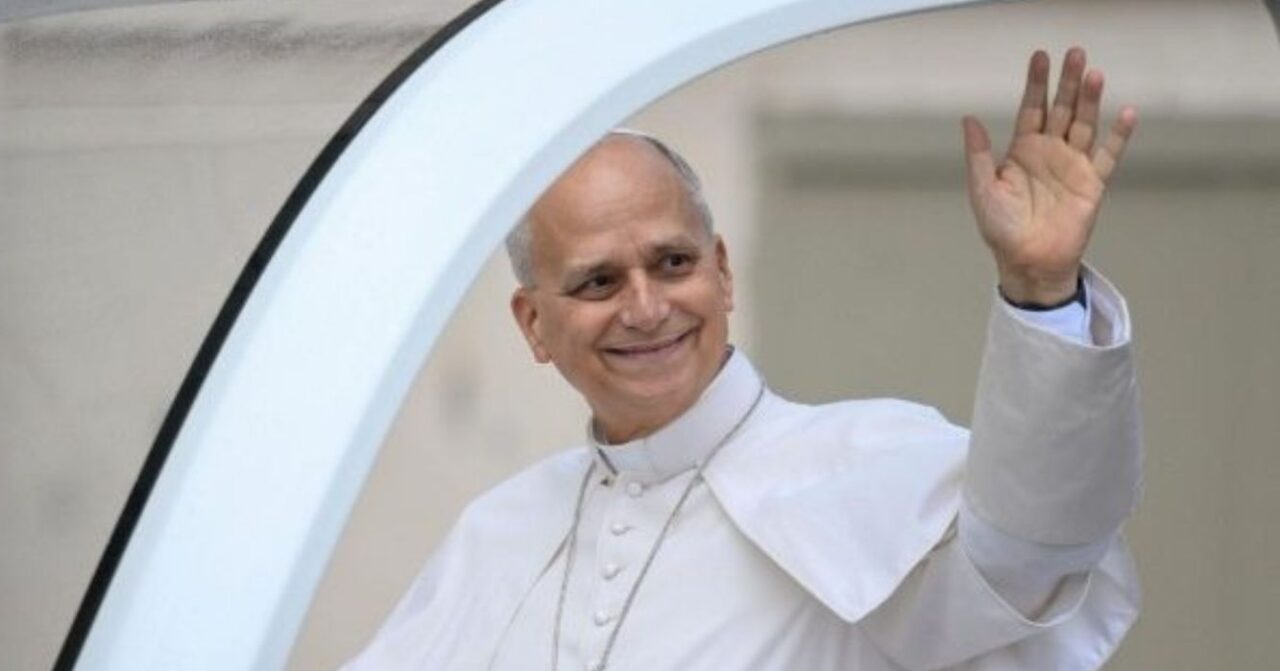 Papa Leão XIV, vestido de branco, sorri e acena com a mão direita enquanto percorre a Praça São Pedro a bordo do papamóvel, durante a Audiência Geral no Vaticano.