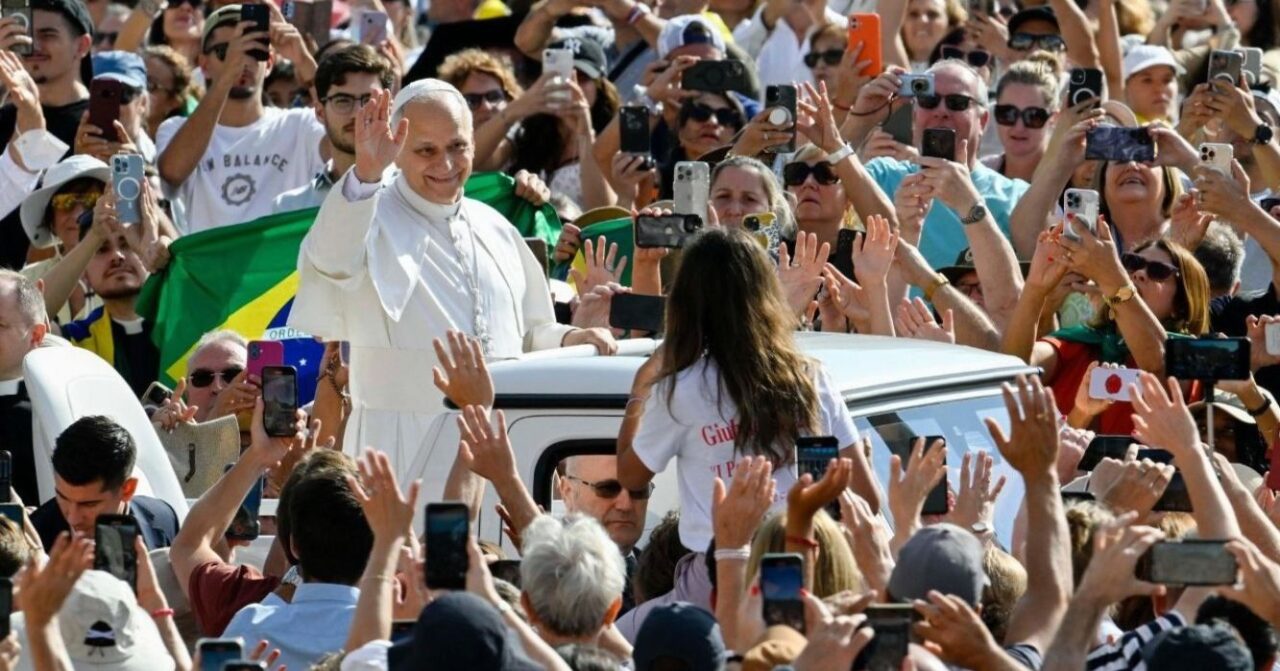 Papa Leão XIV acena para os fiéis durante passagem de papamóvel pela Praça São Pedro, no Vaticano, cercado por uma multidão que o saúda e registra o momento com celulares.