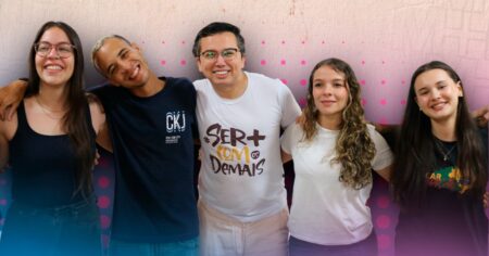 Cinco jovens sorridentes, lado a lado e abraçados, posam para uma foto em frente a um painel com detalhes em rosa. O grupo transmite união, acolhimento e amizade.